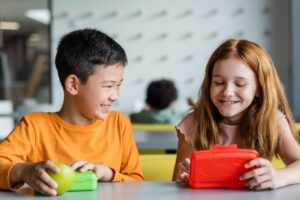 Zwei Kinder sitzen am Tisch und haben eine Brotdose vor sich.
